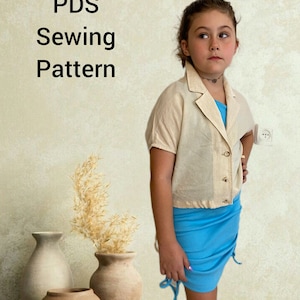 May include: A girl wearing a light beige top with a button closure and a light blue skirt with a drawstring. The skirt has a ruffled hem. The girl is standing in front of a wall with a light beige background. The text "PDS Sewing Pattern" is visible in the top left corner of the image.