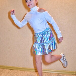 May include: A young person wearing a white long-sleeved shirt with a cut-out shoulder detail and a silver iridescent skirt. They are wearing white sneakers and have their hair pulled back.