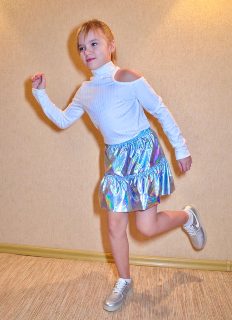 May include: A young person wearing a white long-sleeved shirt with a cut-out shoulder detail and a silver iridescent skirt. They are wearing white sneakers and have their hair pulled back.