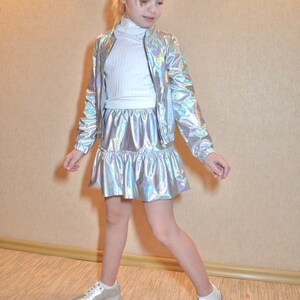 May include: A young person wearing a silver iridescent jacket and skirt set with a white turtleneck top and white sneakers.