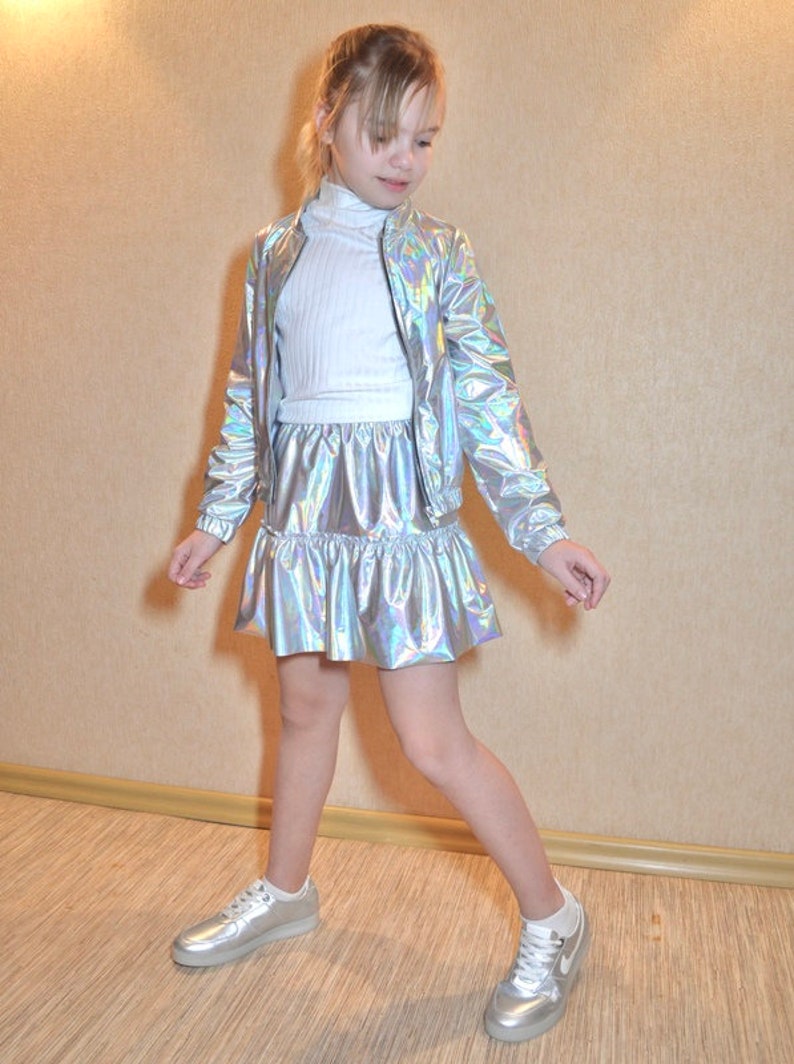 May include: A young person wearing a silver iridescent jacket and skirt set with a white turtleneck top and white sneakers.