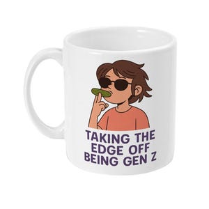 May include: White ceramic mug with a cartoon illustration of a person wearing sunglasses and eating a pickle. The text "TAKING THE EDGE OFF BEING GEN Z" is below the illustration. The mug has a curved handle.