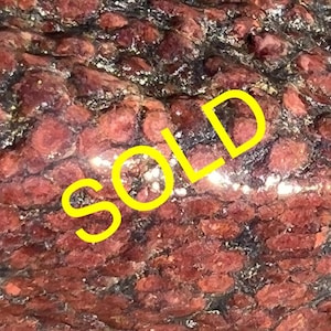 May include: A close-up of a red and black rock with a yellow "SOLD" label over it. The rock has a rough, textured surface and appears to be made of many small crystals.