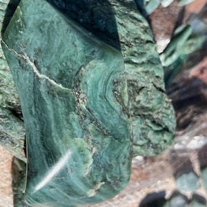 Puede incluir: Primer plano de varias piezas de jade en bruto y pulido. El jade es de un color verde azulado intenso con vetas verdes y negras más oscuras. La piedra tiene una superficie lisa y pulida con una forma natural y orgánica. La imagen muestra la belleza natural de la piedra.