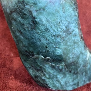May include: A polished, curved stone with a swirling pattern of deep teal and black. The stone has a smooth, rounded top and a textured surface, showcasing natural variations in colour and veining. The stone is set against a red background.