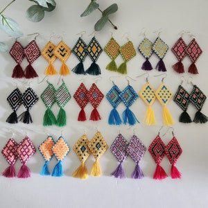 May include: A collection of 24 handmade earrings with colorful geometric patterns and tassels. The earrings are made with embroidery floss and have a variety of colors, including red, blue, green, yellow, and purple.