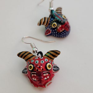 May include: A pair of dangle earrings featuring colorful, hand-painted ceramic animal faces. The earrings are silver-toned and have a hook closure. One earring features a red face with yellow eyes and black horns, while the other earring features a blue face with yellow eyes and black horns.