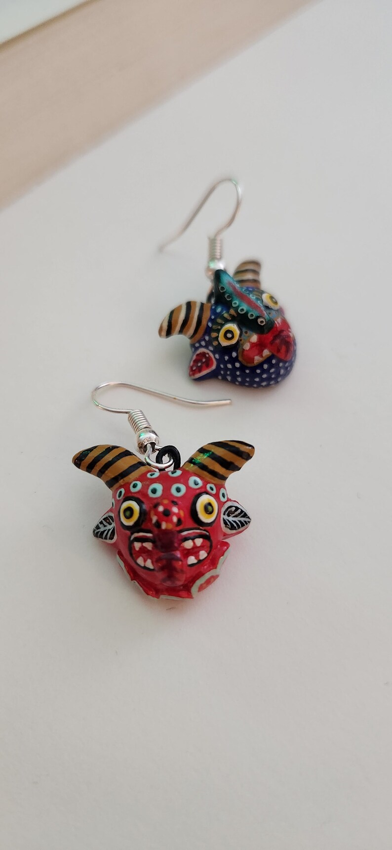 May include: A pair of dangle earrings featuring colorful, hand-painted ceramic animal faces. The earrings are silver-toned and have a hook closure. One earring features a red face with yellow eyes and black horns, while the other earring features a blue face with yellow eyes and black horns.