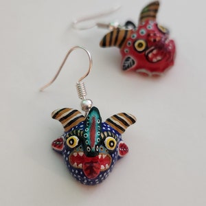 May include: A pair of dangle earrings featuring hand-painted ceramic figures of stylized faces with horns. One face is red with black and white details, the other is blue with black, white, and yellow details.