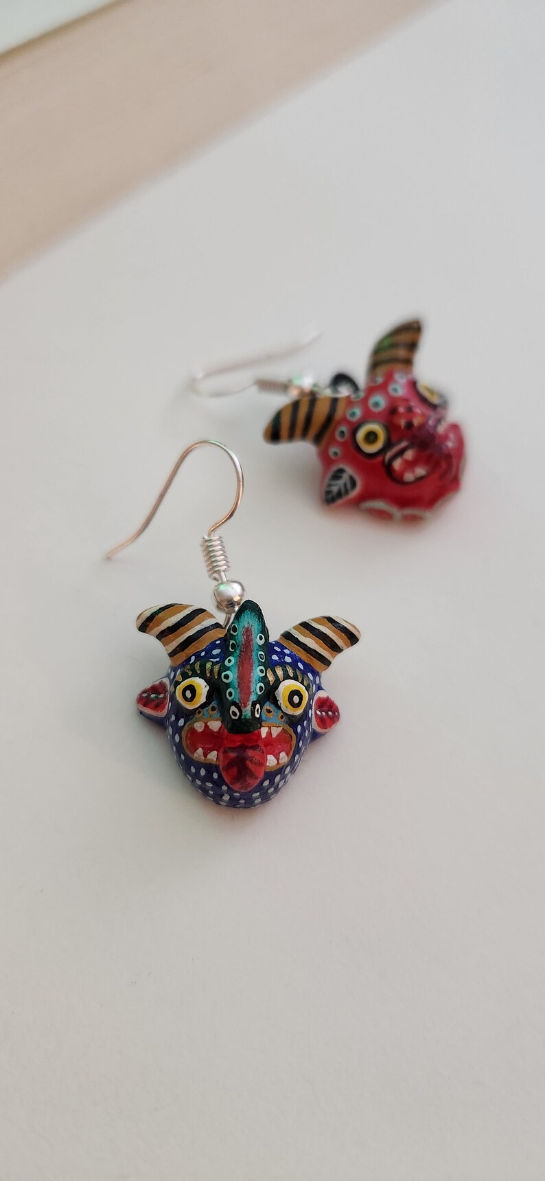 May include: A pair of dangle earrings featuring hand-painted ceramic figures of stylized faces with horns. One face is red with black and white details, the other is blue with black, white, and yellow details.