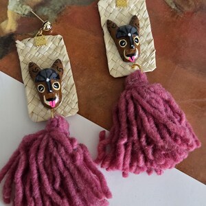 May include: Handmade earrings featuring a dog face design with a pink tongue and a woven background. The earrings have a gold-colored stud and a long, pink tassel. The earrings are a unique accessory.