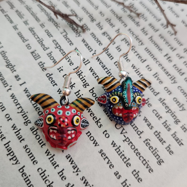 May include: A pair of dangle earrings featuring colorful, hand-painted, ceramic demon masks with silver hooks. The masks have black, red, yellow, blue, and green details.