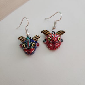 May include: A pair of dangle earrings featuring hand-painted ceramic masks with colorful details. One mask is red with black and white accents, the other is blue with yellow and white accents.