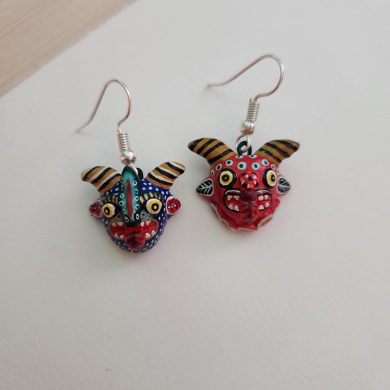 May include: A pair of dangle earrings featuring hand-painted ceramic masks with colorful details. One mask is red with black and white accents, the other is blue with yellow and white accents.
