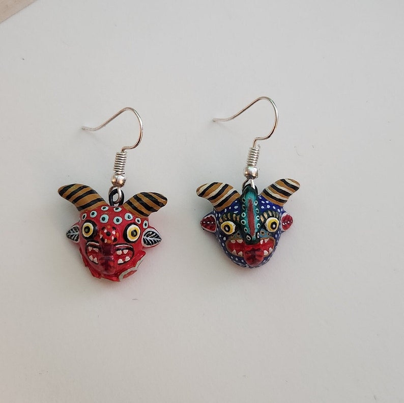 May include: A pair of dangle earrings featuring colorful, hand-painted ceramic masks with intricate details. The masks have a whimsical, almost cartoonish expression with large eyes and a wide open mouth. The earrings are suspended from silver hooks.