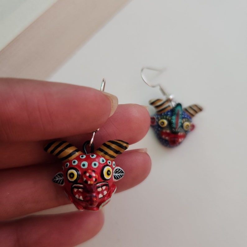 May include: A pair of earrings featuring hand-painted clay figures of colorful, whimsical faces with horns. The figures are red, blue, green, yellow, and black with white accents. The earrings are attached to silver hooks.
