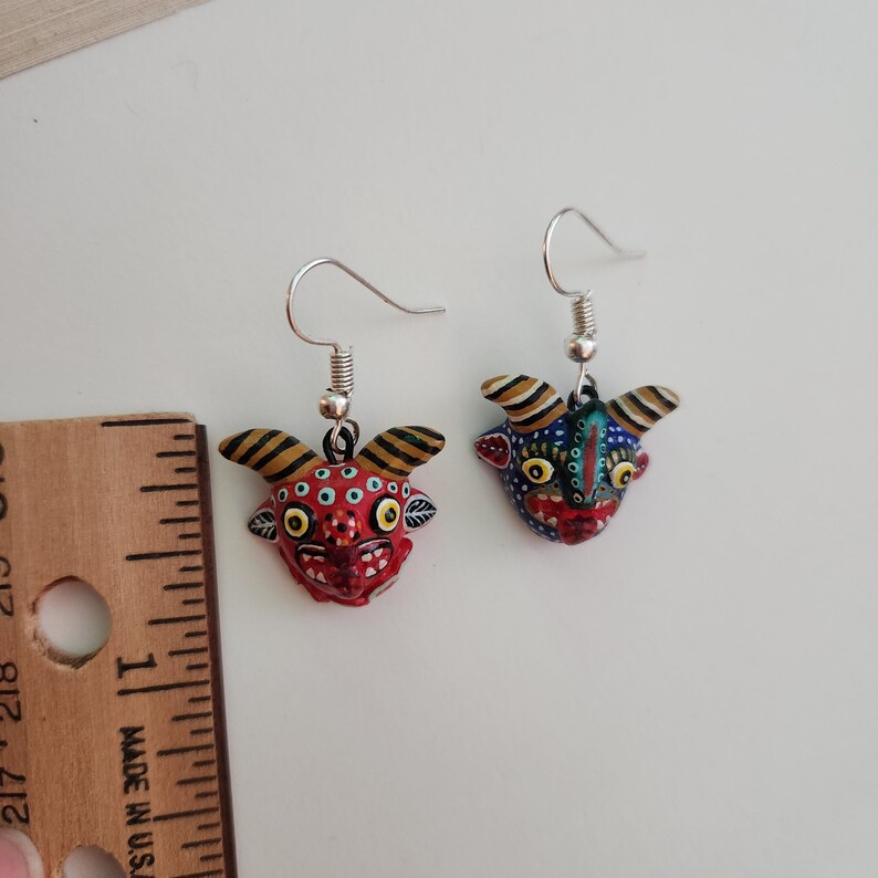 May include: A pair of dangle earrings featuring hand-painted ceramic demon masks. One mask is red with black and yellow accents, the other is blue with green and yellow accents.