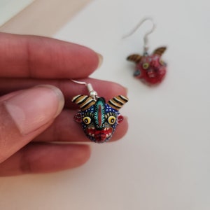 May include: A pair of earrings featuring hand-painted, colorful, detailed masks of a mythical creature with horns. The masks are blue, red, yellow, green, and white with black accents. The earrings are silver.
