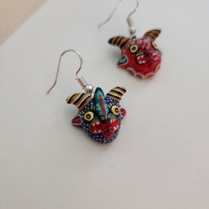 May include: A pair of dangle earrings featuring hand-painted ceramic demon faces. One face is red with black and white details, the other is blue with black, white, and green details.