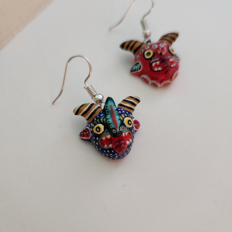 May include: A pair of dangle earrings featuring hand-painted ceramic demon faces. One face is red with black and white details, the other is blue with black, white, and green details.