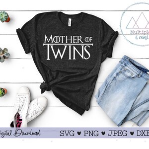 Puede incluir: Una camiseta negra con el texto "Mother of Twins" en letras blancas. La camiseta está sobre una superficie blanca con un par de zapatillas blancas, unos vaqueros azules y una planta en maceta.