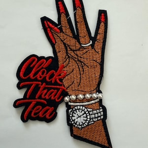 Może przedstawiać: Haftowana naszywka przedstawiająca brązową dłoń z czerwonymi paznokciami, srebrnym pierścionkiem i perłową bransoletką. Widoczny jest również srebrny zegarek. Naszywka zawiera napis "Clock That Tea" w kolorze czerwonym.