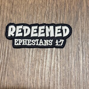 May include: A black and white embroidered patch with the word "REDEEMED" in white block letters above "EPHESIANS 1:7". The patch has a black border and is set against a wood grain background.