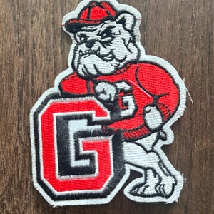 Puede incluir: Un parche bordado que muestra un bulldog blanco con una gorra roja y un suéter rojo. El bulldog se apoya en una letra G grande y negrita, delineada en negro, blanco y rojo. El diseño utiliza hilo de color blanco, negro y rojo en todo.