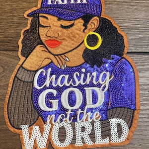 May include: Embroidered patch depicting a Black woman in a purple baseball cap with "FAITH" and a sequined top. The text "Chasing GOD not the WORLD" is in white. The patch has a brown border. The image is on a wooden surface.