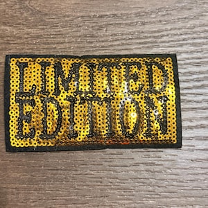 May include: A rectangular patch with the words "LIMITED EDITION" in black letters. The letters are on a gold sequined background, with a black border. The patch is on a wooden surface.