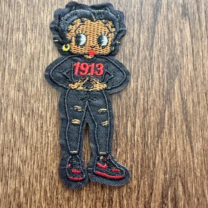 May include: Embroidered patch featuring a cartoon character in black denim jeans and a black top with the number "1913" in red. The character has brown skin, black hair, and red sneakers. The patch is on a wooden surface.