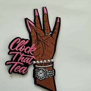 May include: Embroidered patch featuring a brown hand with pink fingernails, a silver ring, and a watch. The patch includes the phrase "Clock That Tea" in pink and black lettering. The hand is adorned with a pearl bracelet.