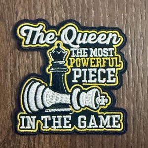 Może przedstawiać: Czarno-biało-żółta naszywka z haftem przedstawiająca figurę szachowej królowej oraz tekst: The Queen THE MOST POWERFUL PIECE IN THE GAME. Wzór zawiera pionową czarną królową i poziomą białą figurę szachową z żółtymi obwódkami.