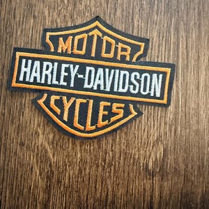 May include: A black and orange Harley-Davidson Cycles patch. The patch features the Harley-Davidson logo with the words "MOTOR HARLEY-DAVIDSON CYCLES" in white and orange lettering. The patch is rectangular with a shield-like shape.