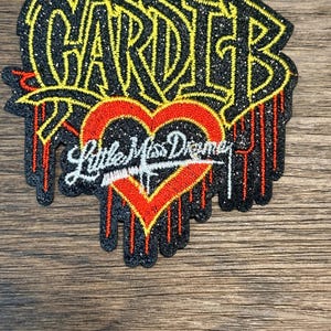Könnte beinhalten: Gestickter Aufnäher mit dem Namen "Cardi B" in Gelb, umrandet von Schwarz, mit einem roten Herz und dem Text "Little Miss Dreame" in Weiß. Der Aufnäher hat einen glitzernden schwarzen Hintergrund und rote Tropfenakzente.