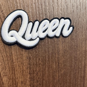 May include: A white embroidered patch featuring the word Queen in a cursive script font. The lettering has a thick, raised texture and is outlined with a bold black border, creating a high-contrast design suitable for application onto fabric.