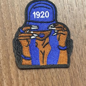May include: An embroidered patch featuring a figure with brown skin wearing a blue baseball cap with the white text 1920. The figure has long dark hair, blue clothing accents, and long white-tipped fingernails, all outlined in a black border.