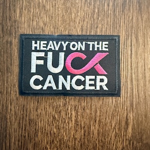 May include: Black rectangular patch with white text that reads "HEAVY ON THE FUCK CANCER". The word "FUCK" is in white with a pink ribbon replacing the "U". The patch is on a wooden surface.