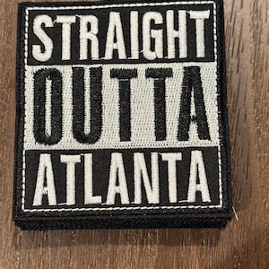 May include: Black and white embroidered patch with the text "STRAIGHT OUTTA ATLANTA". The patch has a rectangular shape with a white border and is likely designed to be sewn or ironed onto clothing or accessories.