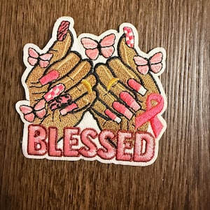 May include: Embroidered patch featuring two hands with manicured nails in shades of pink and gold, adorned with pink butterflies. The word "BLESSED" is embroidered in pink letters, and a pink ribbon is also present.