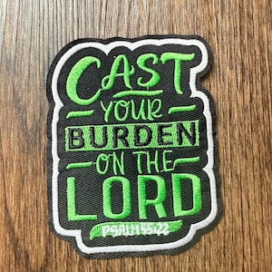 May include: Embroidered patch with the text "CAST YOUR BURDEN ON THE LORD" in green script and block letters on a black background. The patch has a white border and the text "PSALM 55:22" at the bottom. The patch is rectangular with rounded corners.