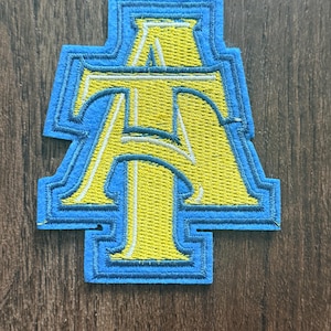 May include: An embroidered patch featuring the letters A and T intertwined in yellow thread with a white outline. The design is bordered by a thick, light blue felt-like material that follows the shape of the letters, creating a layered appearance.