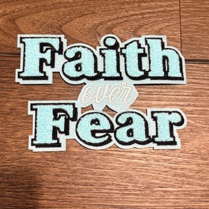 Puede incluir: Dos parches bordados con las palabras "Faith over Fear". El texto es de color azul claro con un contorno negro y un fondo blanco. La palabra "over" está en una fuente cursiva más pequeña.