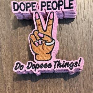 May include: Pink patch with the words "DOPE PEOPLE" stacked above a hand making a peace sign. The hand is tan with pink fingernails and a silver ring. Below the hand is the phrase "Do Dopeee Things!"