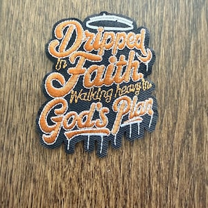 Puede incluir: Parche bordado con las palabras "Dripped in Faith Walking heavy in God's Plan" en escritura naranja sobre un fondo negro. El parche tiene un borde blanco y un diseño de halo.