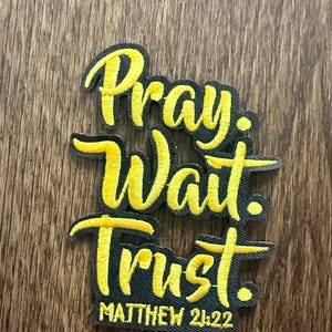 May include: Embroidered patch with the words "Pray. Wait. Trust." in yellow script on a black background. The text "Matthew 21:22" is below. The patch is on a wooden surface.