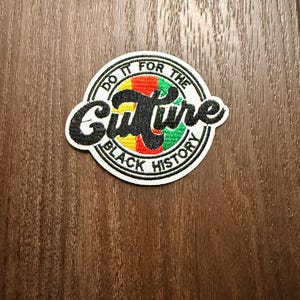 Può includere: Una toppa circolare con le parole "Do it for the Culture" e "Black History" in un carattere nero stilizzato. La parola "Culture" è in un design colorato di rosso, giallo e verde. La toppa ha un bordo bianco.