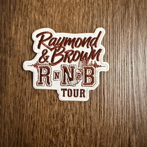 Könnte beinhalten: Ein weißer Aufnäher mit braun gesticktem Text, der "Raymond & Brown RNB TOUR" lautet. Der Aufnäher hat eine dekorative Bordüre und eine Mikrofon-Grafik. Er liegt auf einer Holzoberfläche.