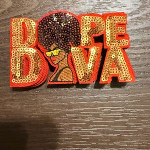 May include: A red and gold sequined patch with the words "DOPE DIVA" and an illustration of a woman with sunglasses and a large afro. The patch is on a wooden surface.