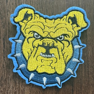 May include: An embroidered patch featuring a yellow bulldog head with a stern expression, wearing a dark blue spiked collar. The design is outlined in light blue thread, showcasing detailed stitching of the dog's facial features and silver spikes.